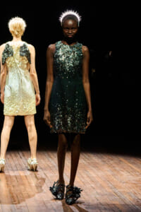 Black model on a fashion runway wearing a teal sequined mini dress with a spiked crown headpiece, center stage with another model in the background.
