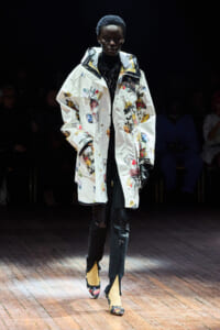 Model on a runway wearing a long white butterfly-printed coat over a black outfit, with jeweled boots and gloves in a dim setting.
