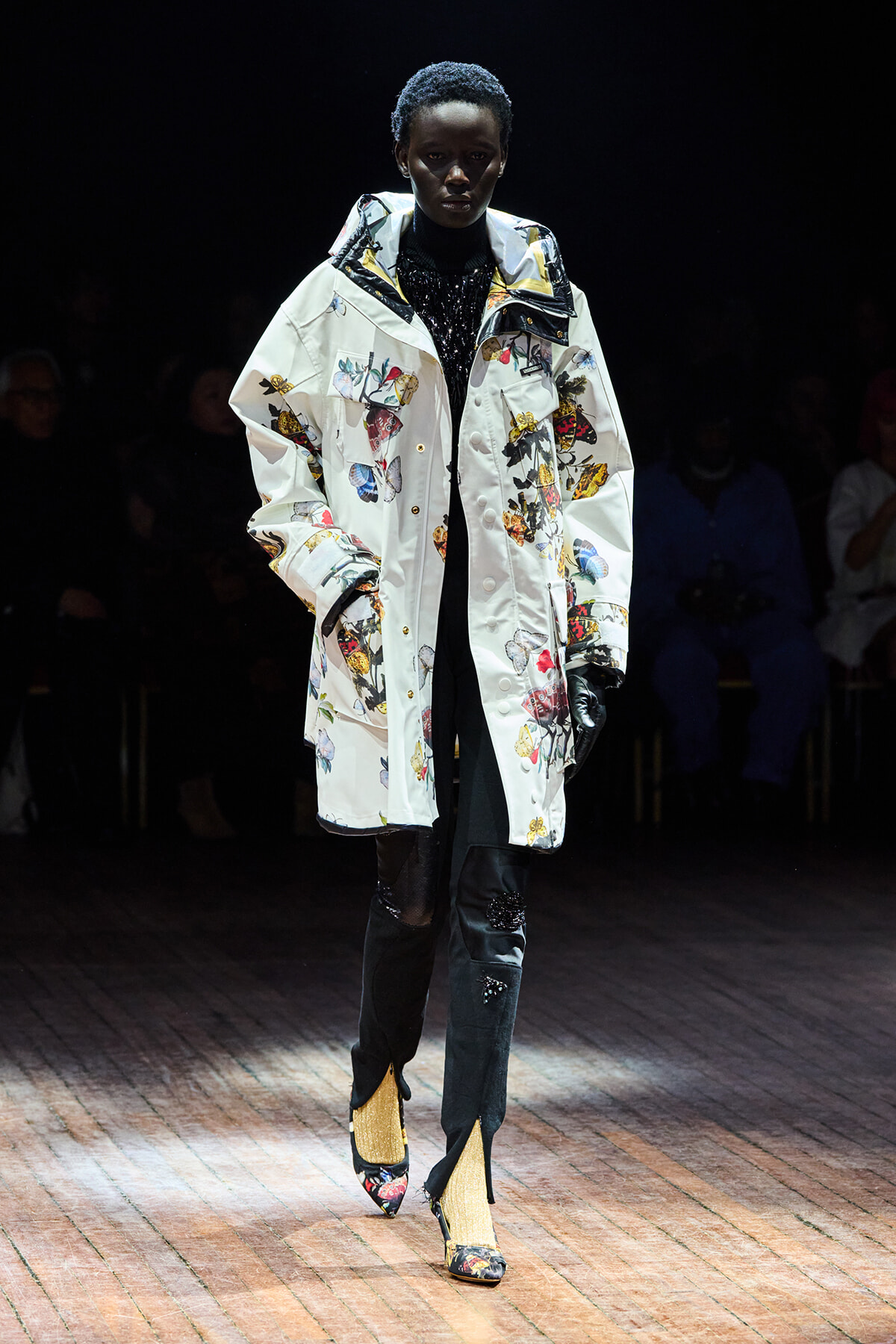 Model on a runway wearing a long white butterfly-printed coat over a black outfit, with jeweled boots and gloves in a dim setting.
