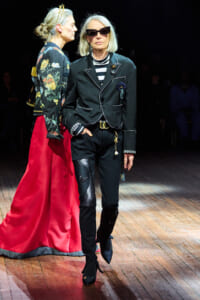 Two women walk a fashion runway: one in a black tailored suit with white piping and sunglasses, the other in a floral jacket and bright red skirt.