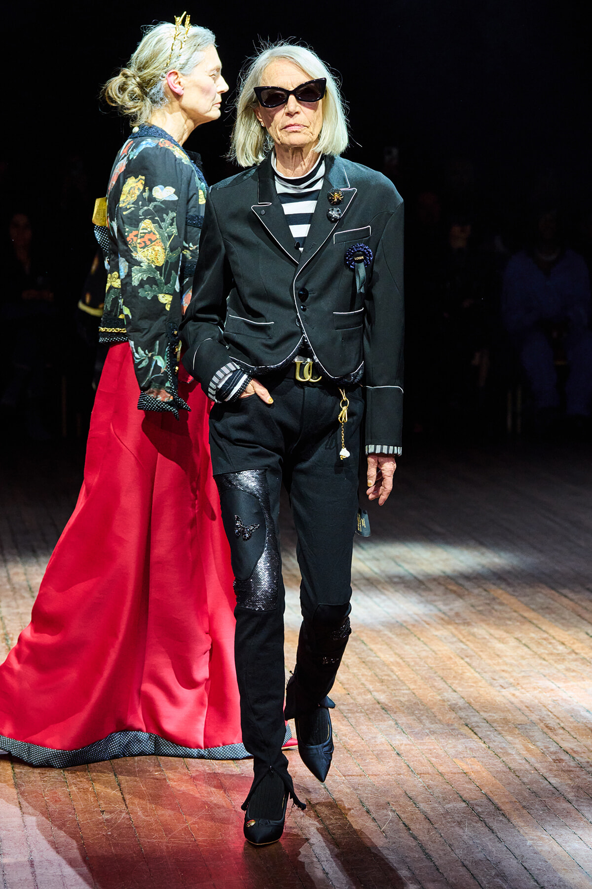 Two women walk a fashion runway: one in a black tailored suit with white piping and sunglasses, the other in a floral jacket and bright red skirt.