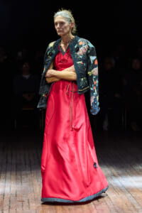 Older woman wearing a red satin gown with a floral-patterned jacket and a small crown, walking a runway.
