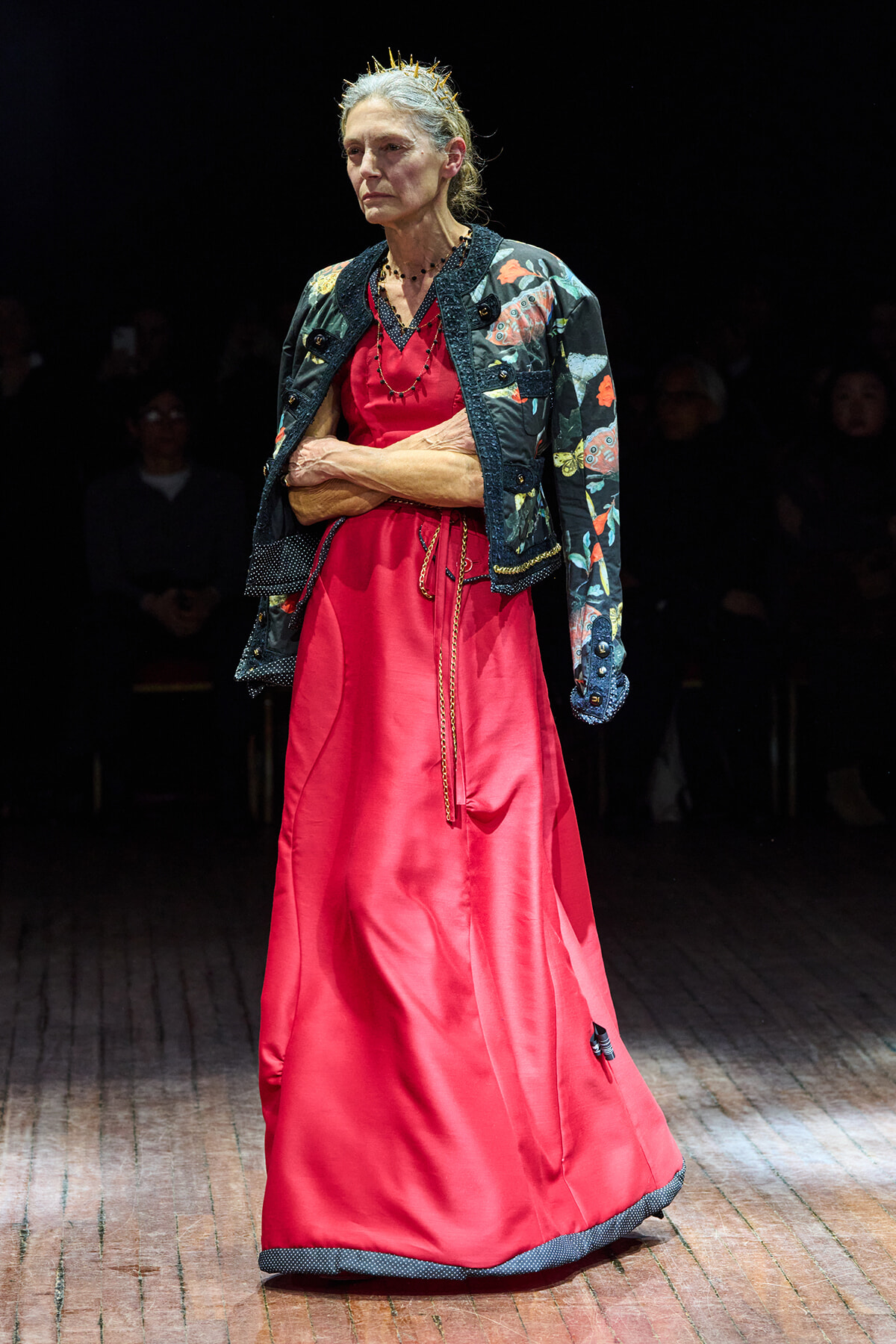 Older woman wearing a red satin gown with a floral-patterned jacket and a small crown, walking a runway.