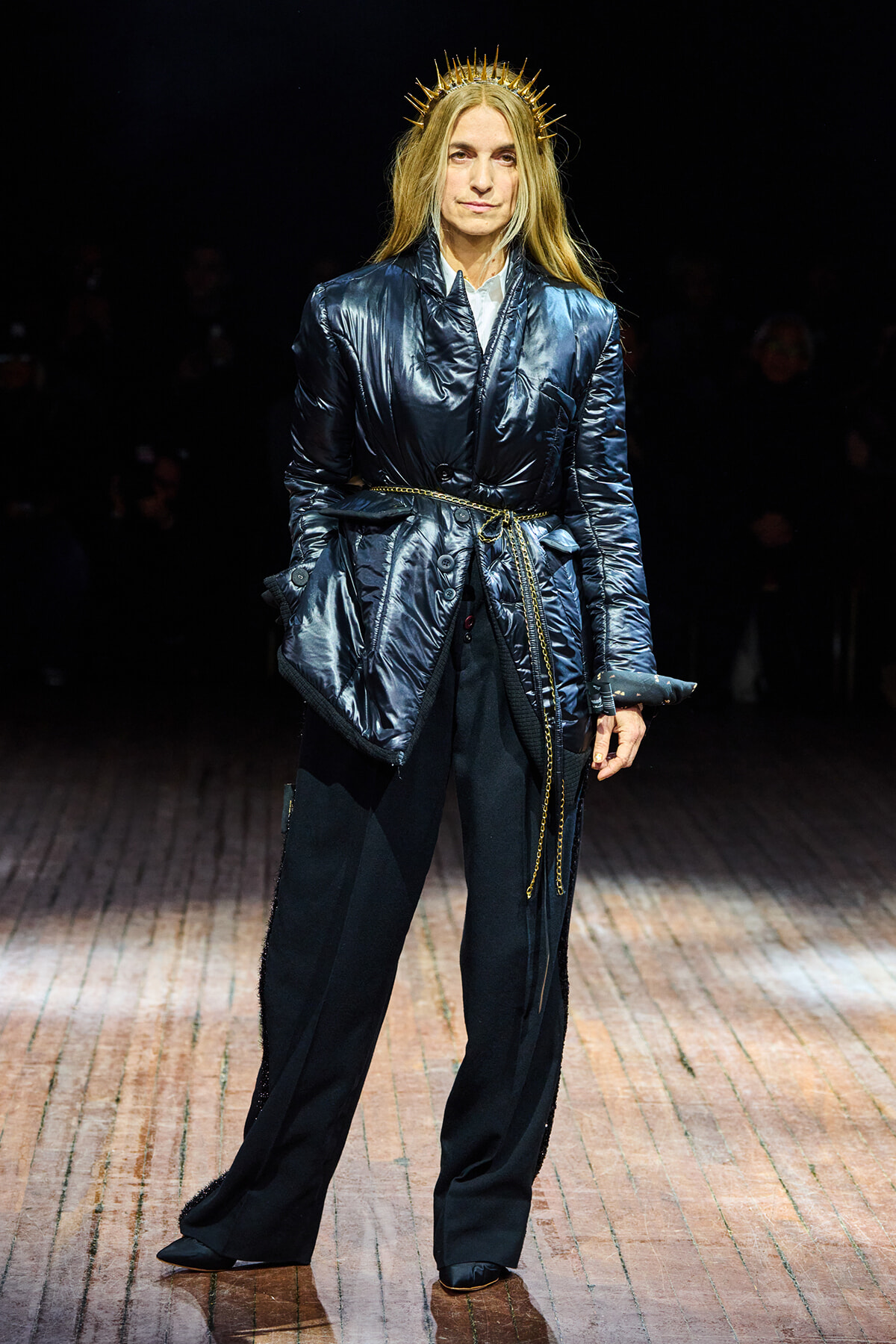 Model on a runway wearing a glossy navy quilted coat belted with a rope, paired with black wide-leg pants and a gold spiked halo crown.