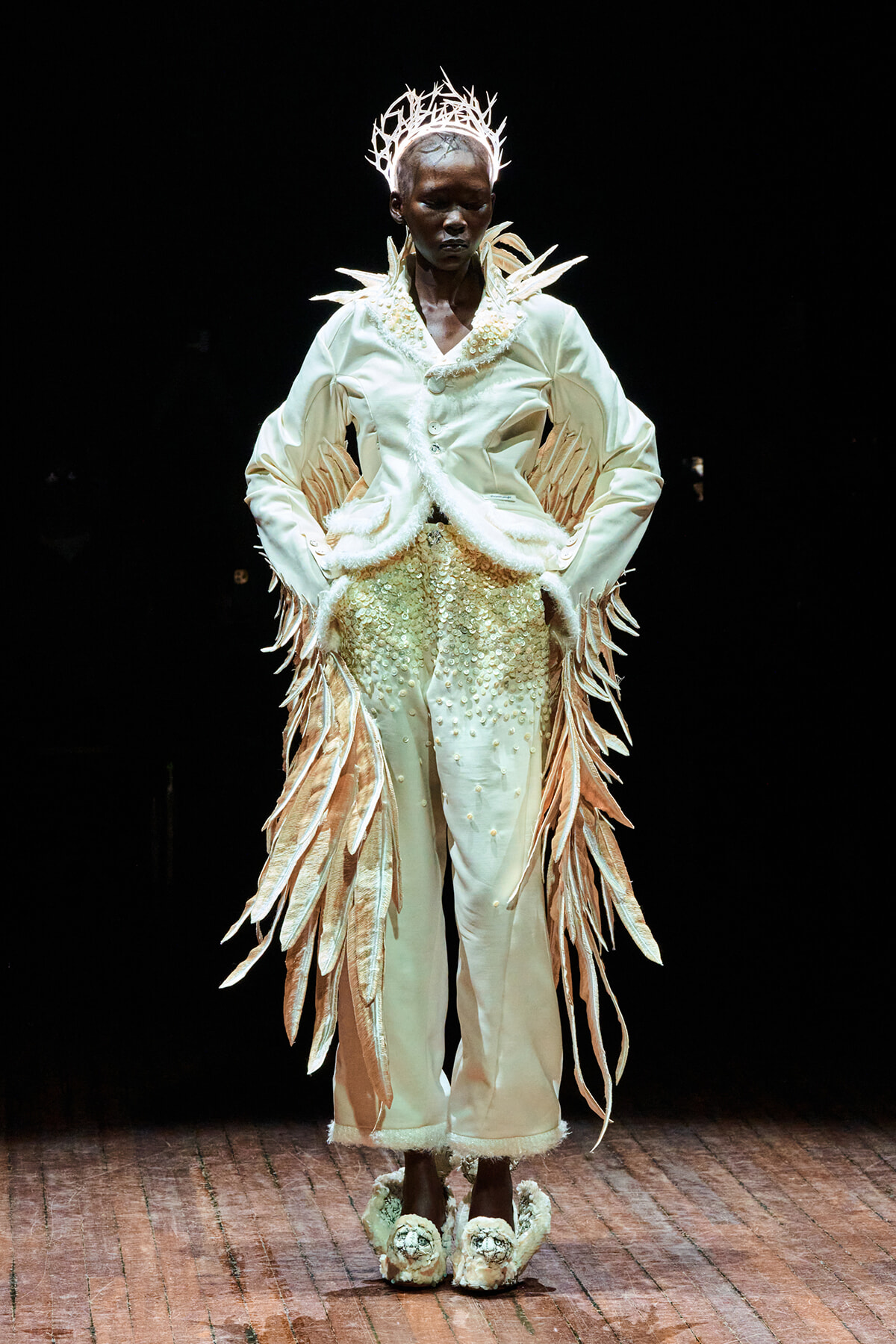 Model on a runway wearing an ivory avant-garde outfit with feather-like fringe, pearl details, and a crown-shaped headpiece.