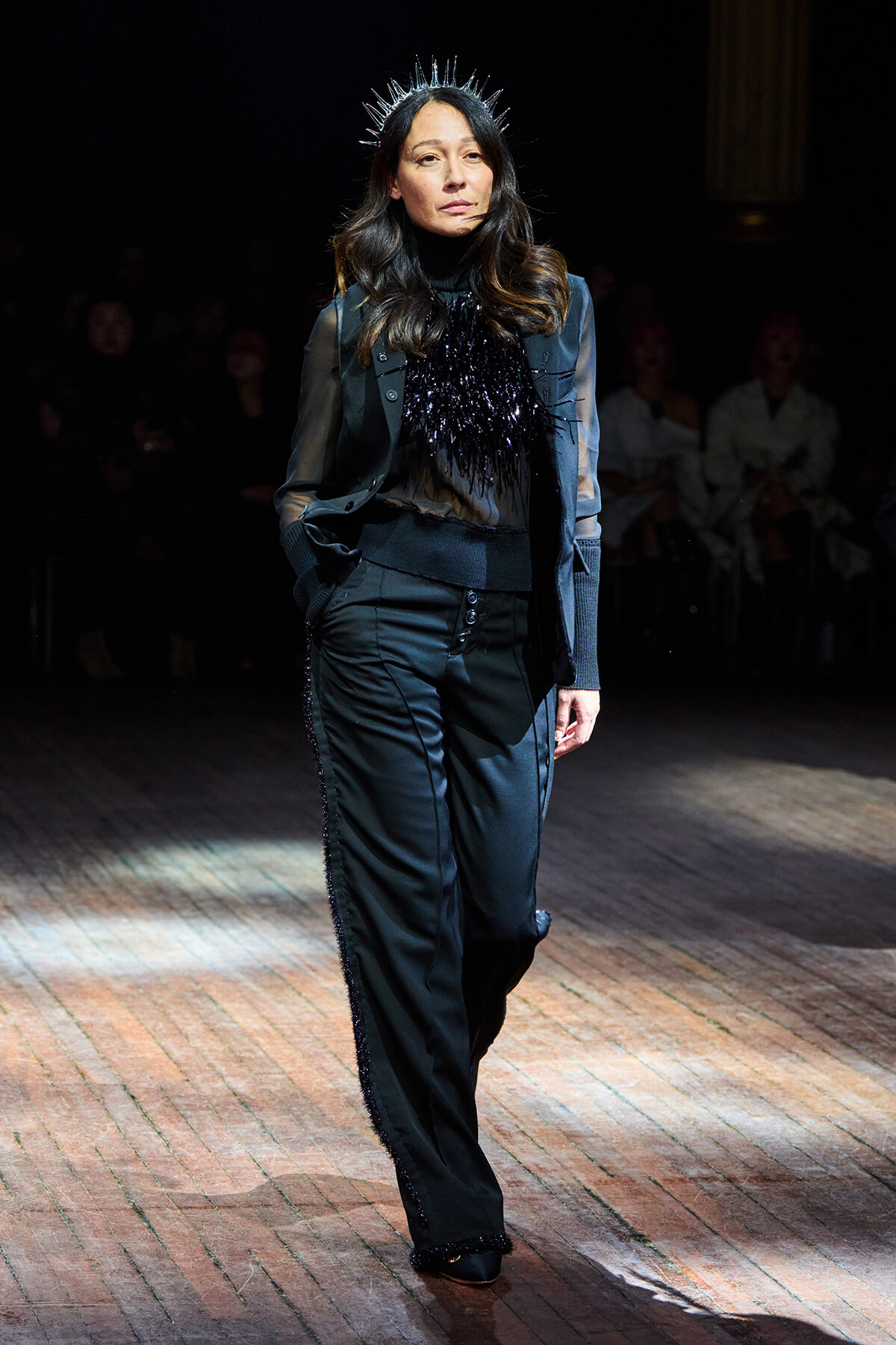 Model walks the runway in a black suit with sheer sleeves and a feathered bib, wearing a spiked silver crown.