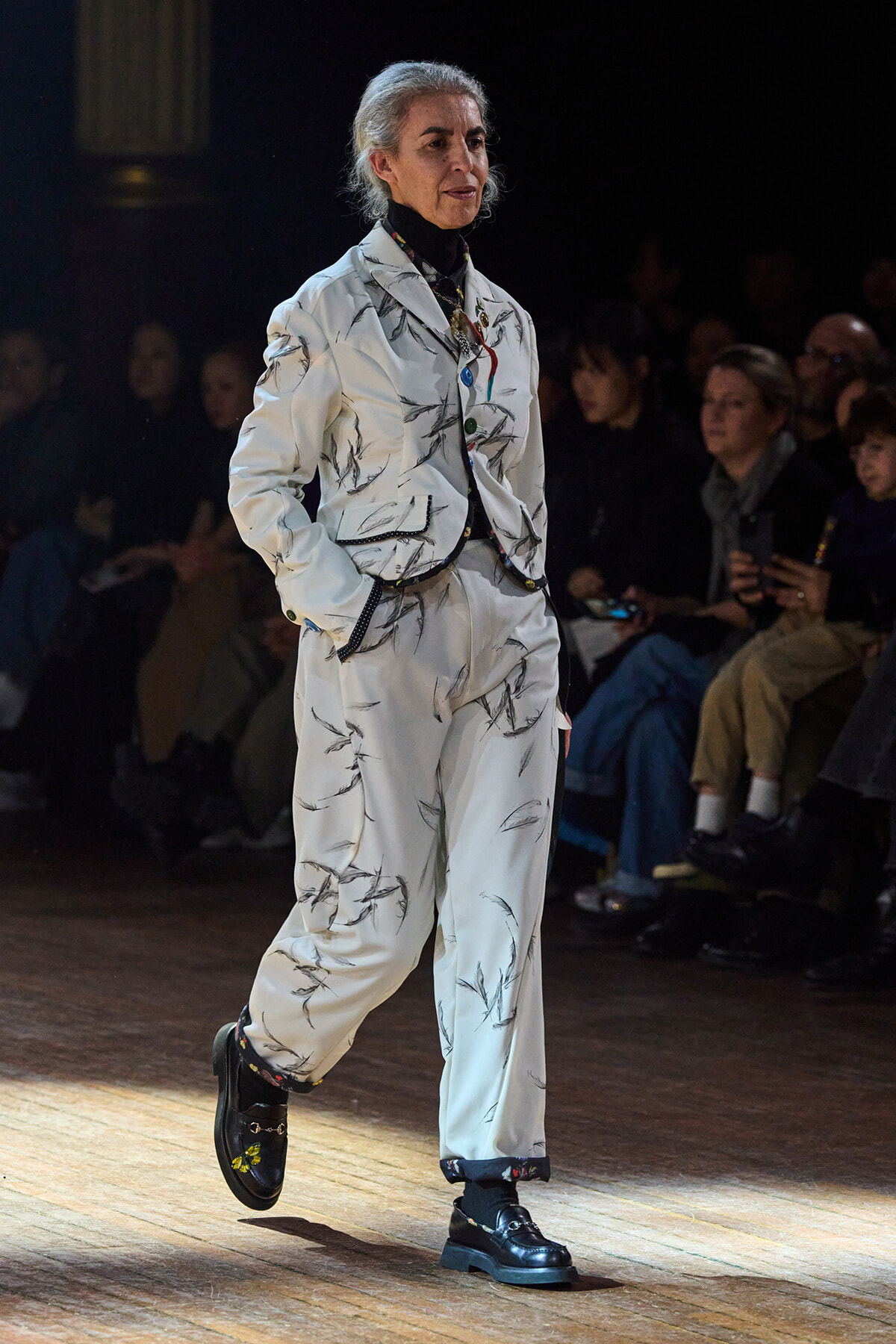 Female model walking the runway in a white blazer and matching pants with black brush-stroke pattern, black loafers, audience in the background.