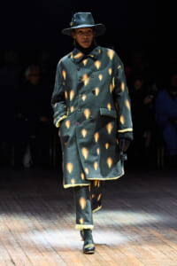 Model walking a runway in a teal double-breasted coat with orange flame-like prints, a wide-brim hat, and gloves.
