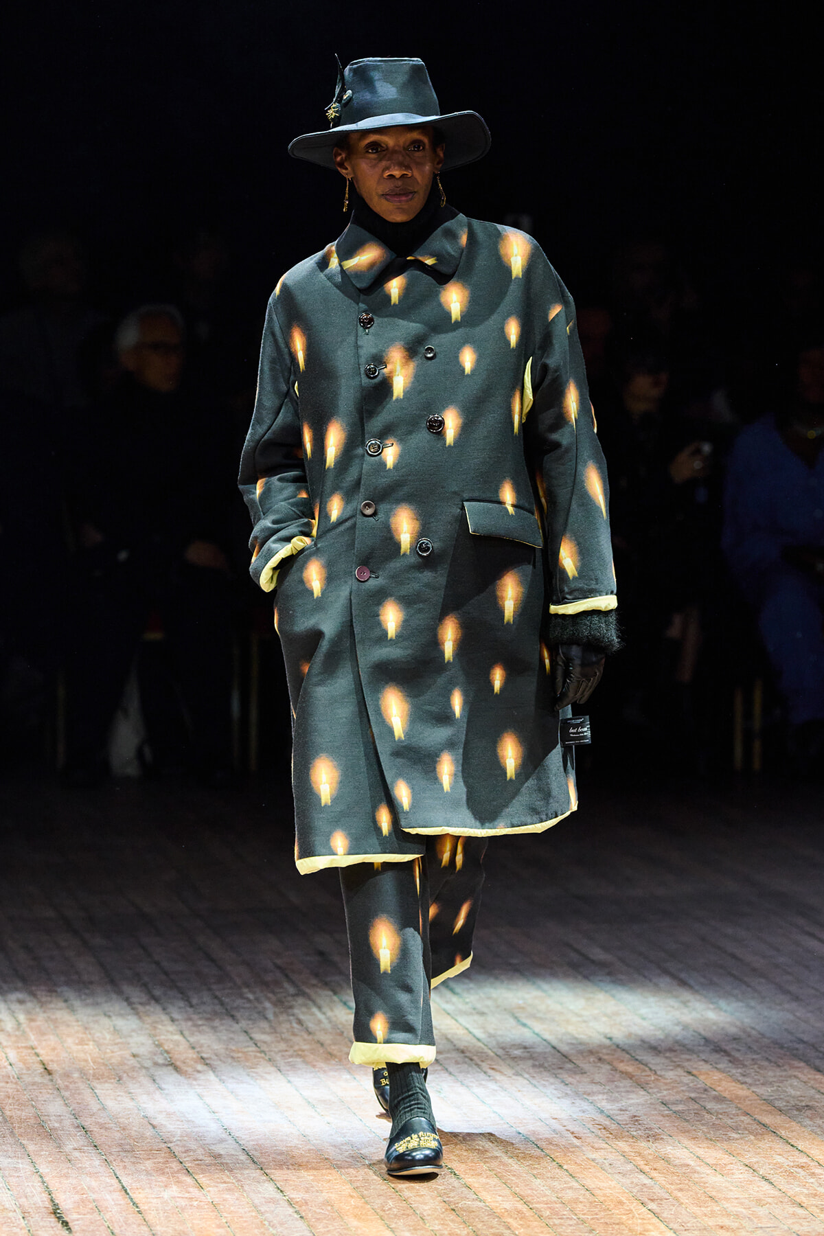 Model walking a runway in a teal double-breasted coat with orange flame-like prints, a wide-brim hat, and gloves.