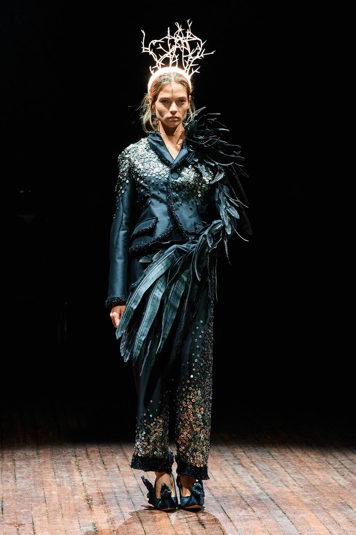 Model walking a runway in a teal sequined suit with sculptural feather details and a white branch headpiece against a dark background.