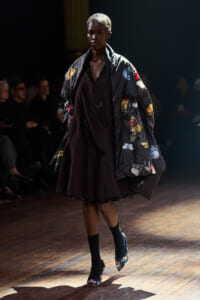 Model walks the runway in a dark brown dress topped with a large floral-embroidered coat on a dimly lit stage.