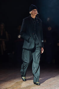 Model walking a runway in a dark tailored suit with a knit beanie and subtle embellishments on the hems and pockets.