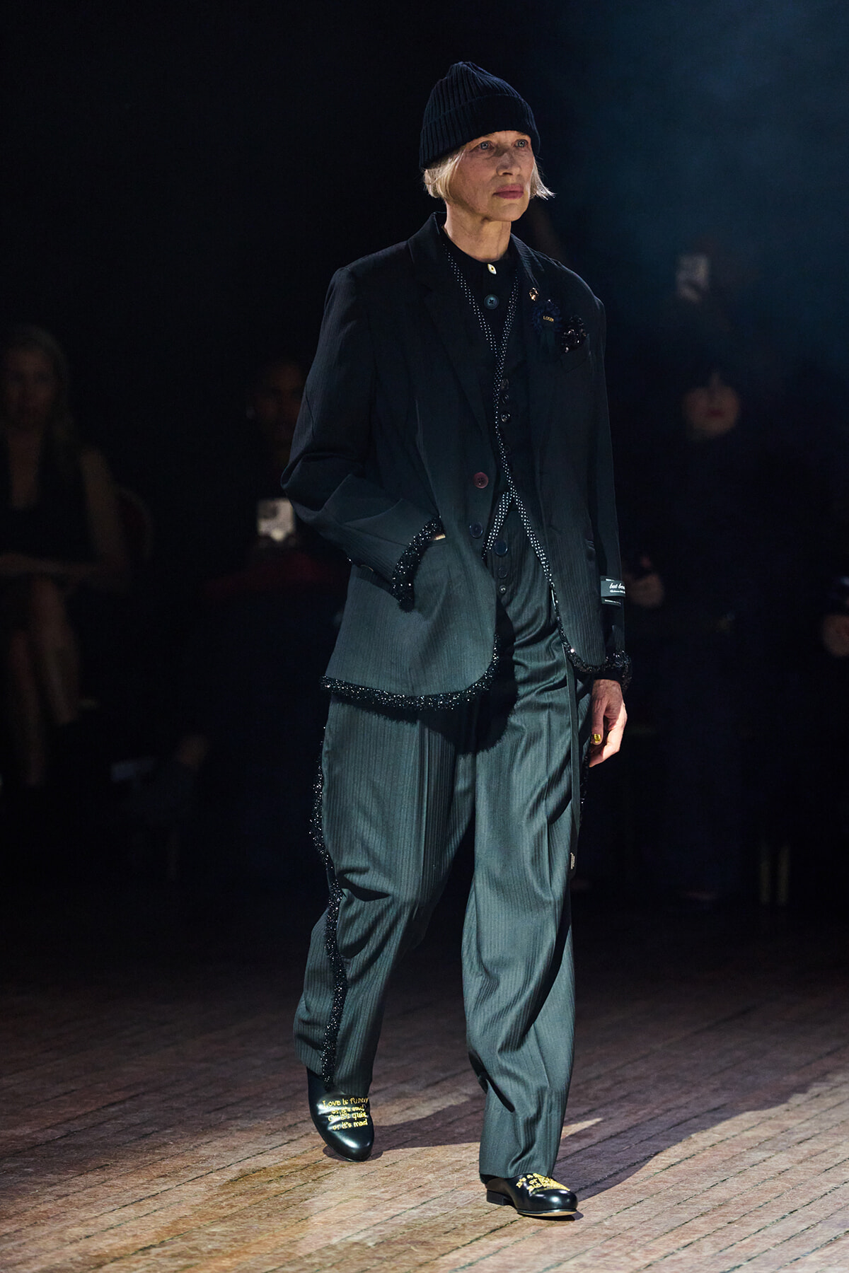 Model walking a runway in a dark tailored suit with a knit beanie and subtle embellishments on the hems and pockets.