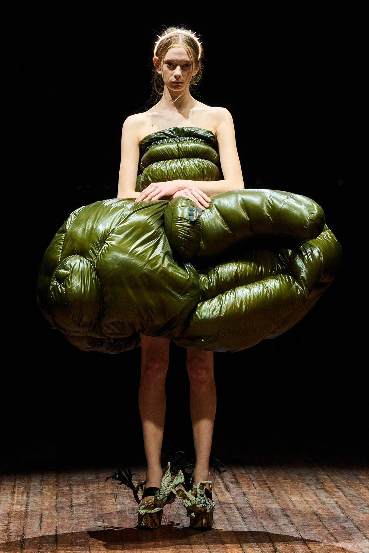 Model on a dark stage wearing a large shiny olive-green inflatable dress, hands clasped in front on a wooden runway.