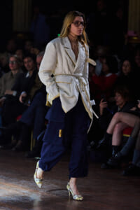 Female model walks a runway wearing a cream belted blazer over navy cargo pants and floral heels, with a seated audience in the background.