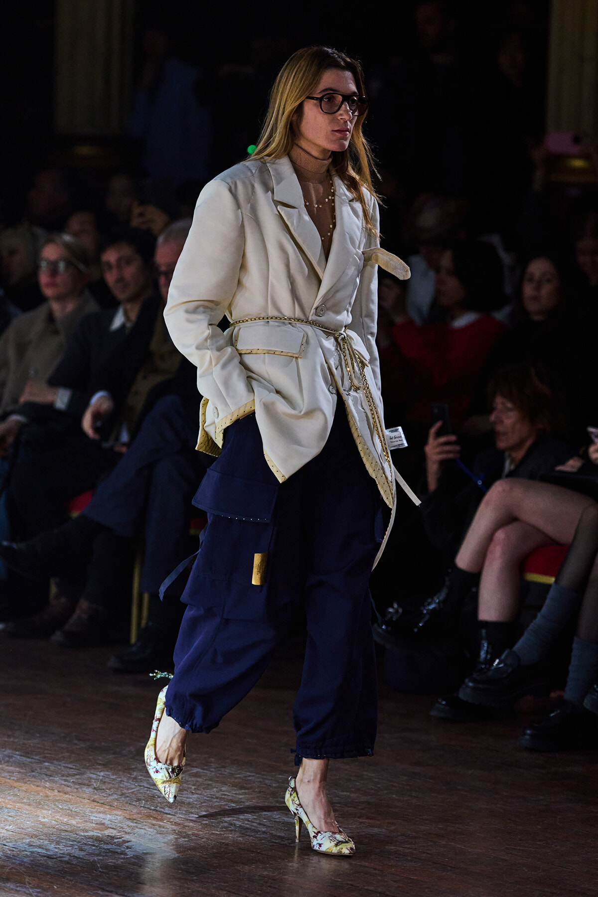 Female model walks a runway wearing a cream belted blazer over navy cargo pants and floral heels, with a seated audience in the background.