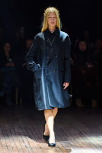 Model walking a fashion show in a dark blue oversized coat with a gold feather pendant necklace and a spiked crown headpiece, spotlighted on the runway.