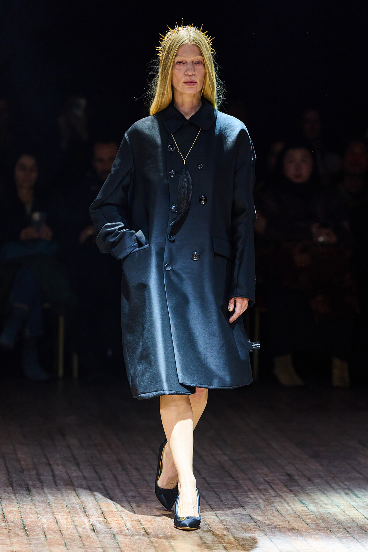 Model walking a fashion show in a dark blue oversized coat with a gold feather pendant necklace and a spiked crown headpiece, spotlighted on the runway.