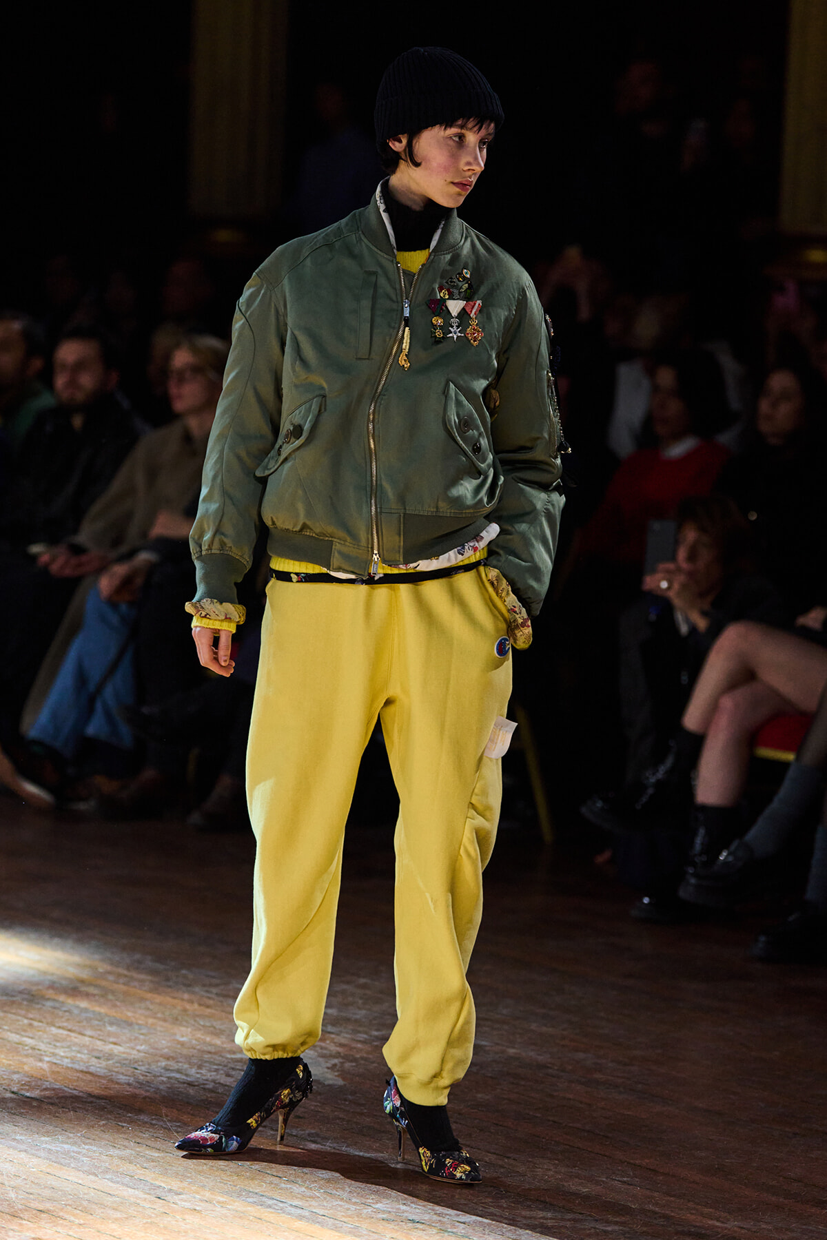Model on a runway wearing an olive bomber jacket with patches and yellow jogger-style pants, black beanie, floral heels.