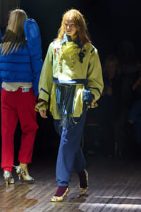 Model walking a fashion runway in a pale yellow jacket with gold buttons, blue belt, fringe skirt, and patterned heels as audience looks on.