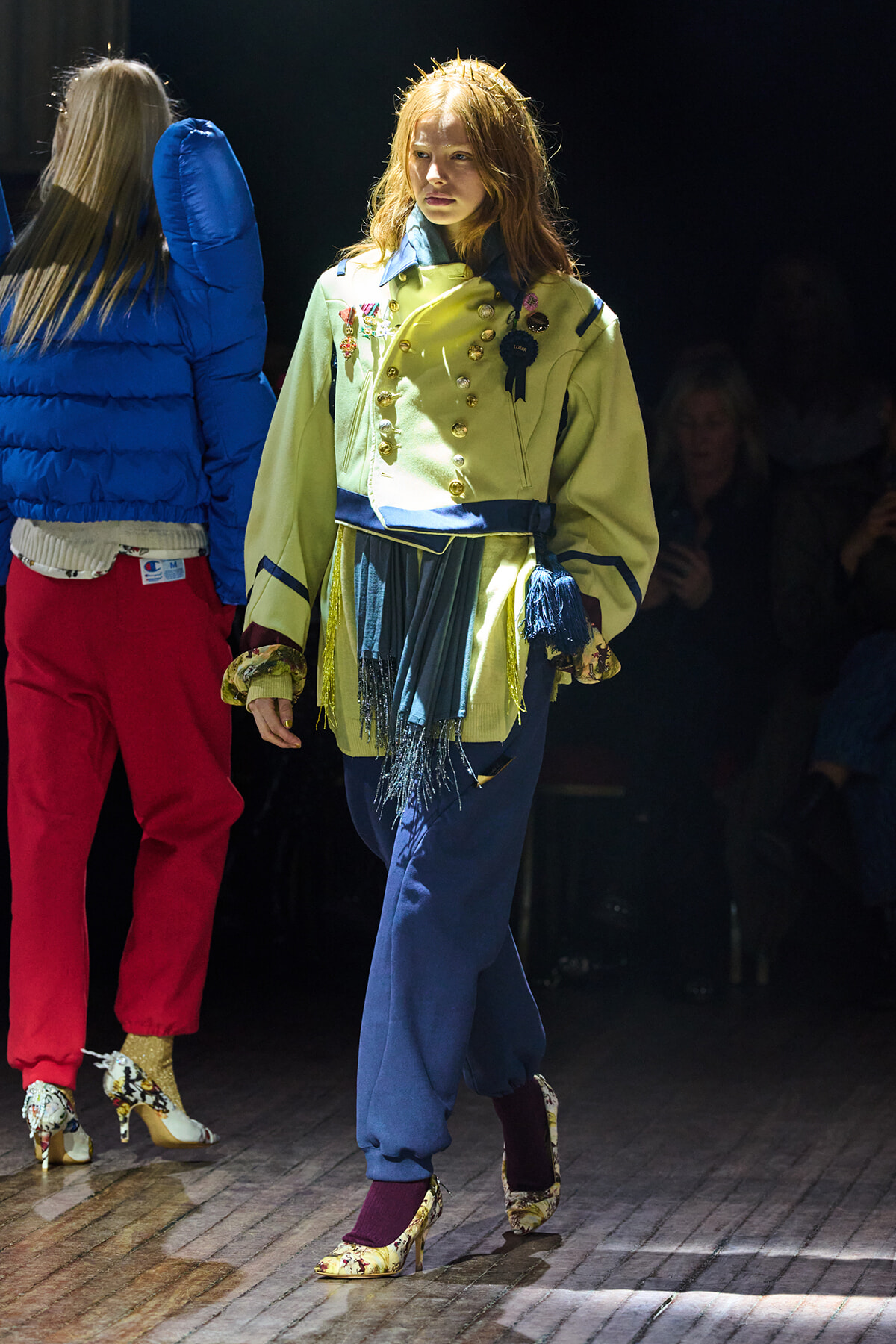 Model walking a fashion runway in a pale yellow jacket with gold buttons, blue belt, fringe skirt, and patterned heels as audience looks on.