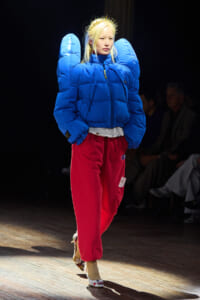 Model walks a runway in an oversized bright blue puffer jacket, red sweatpants, floral heels, and a gold crown headpiece on a lit catwalk.