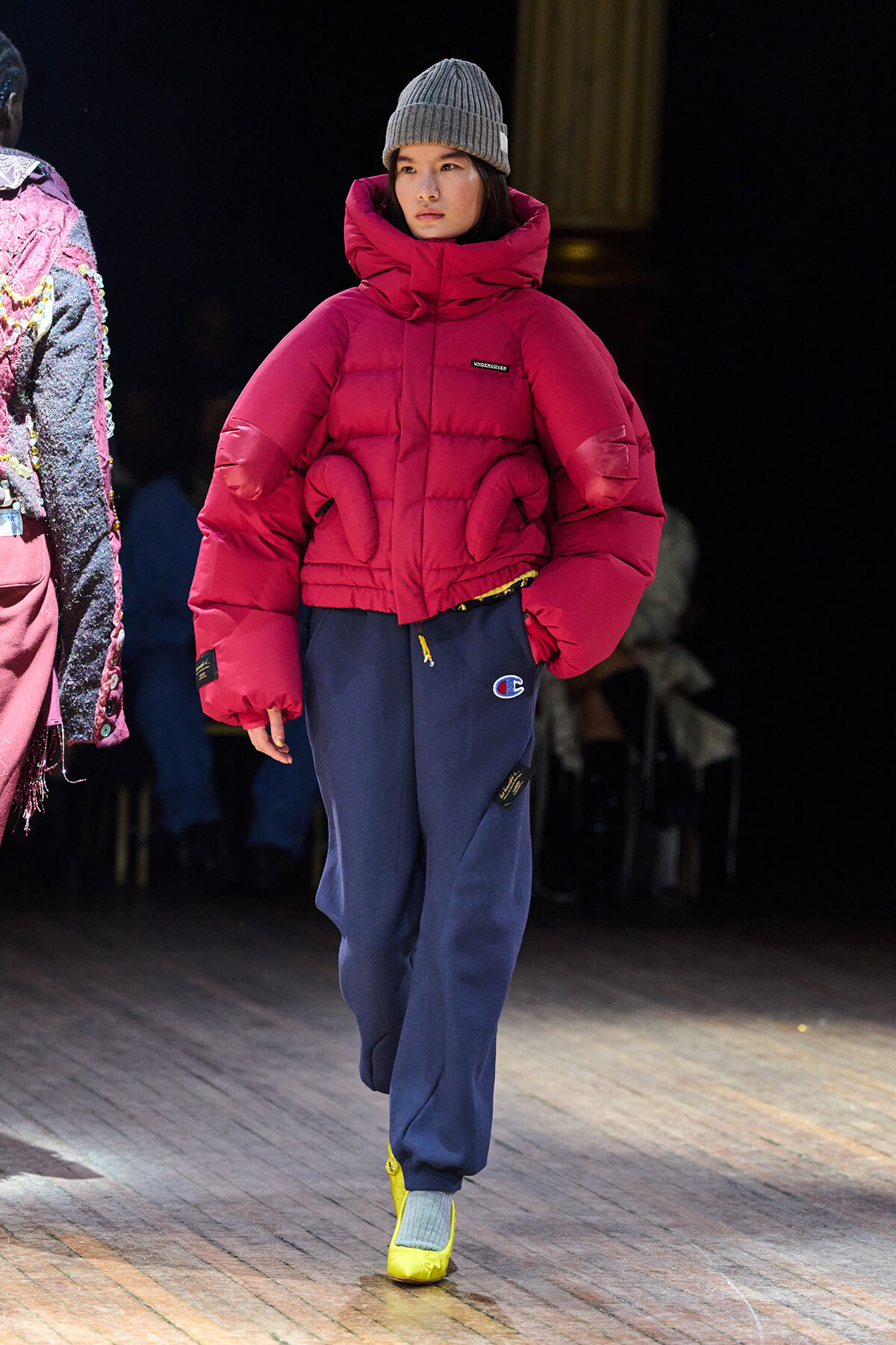 Runway model wearing a bright red oversized puffer jacket with a large hood, gray knit beanie, navy sweatpants, and yellow sneakers.
