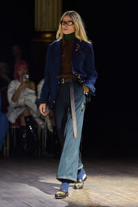 Model walking a fashion show runway in a blue velvet blazer, brown turtleneck, wide teal jeans, and loafers.
