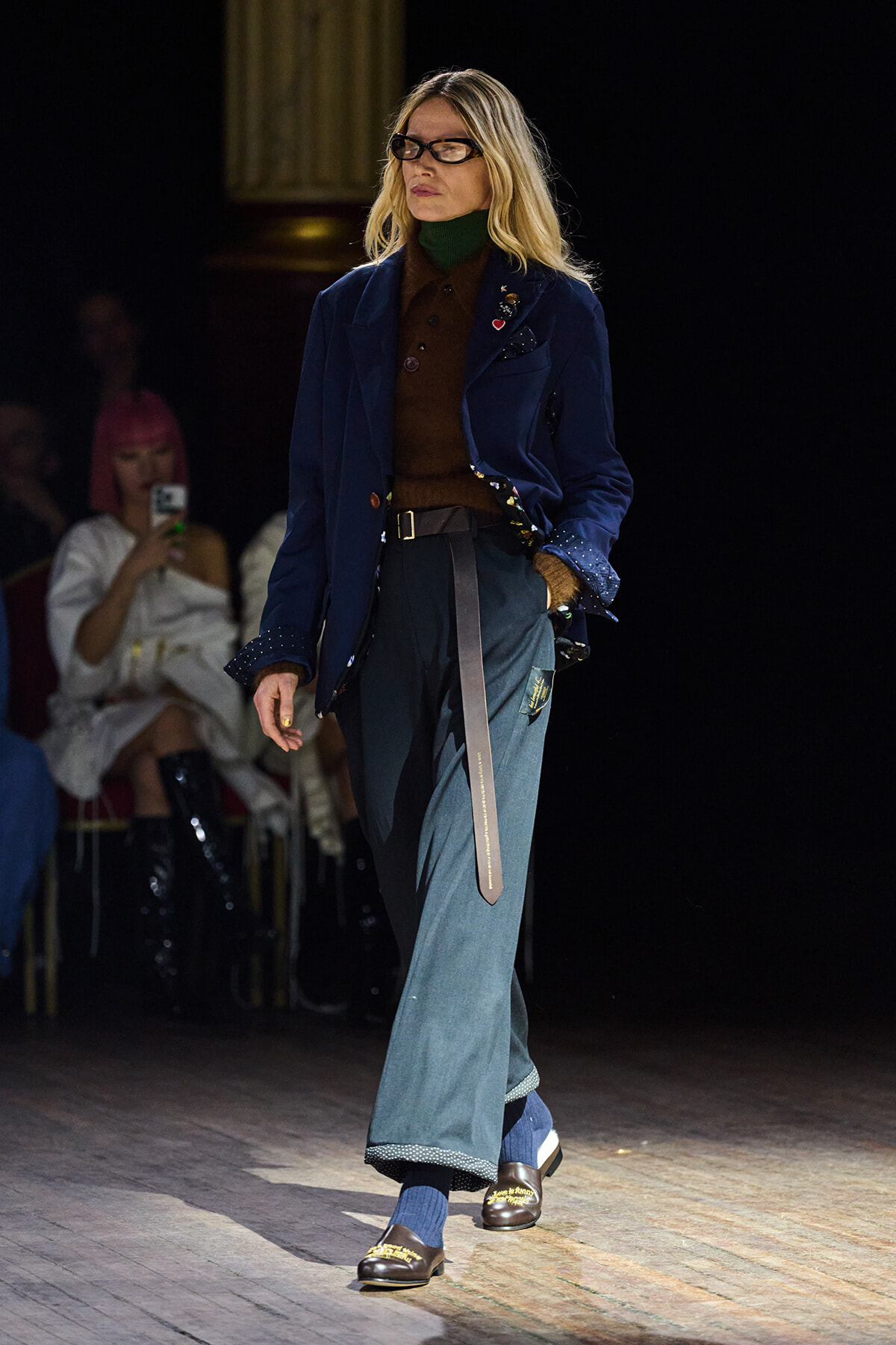 Model walking a fashion show runway in a blue velvet blazer, brown turtleneck, wide teal jeans, and loafers.
