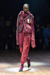 Model walks a fashion show in a burgundy patchwork quilted coat with beads and fringe, paired with matching pants and heels.