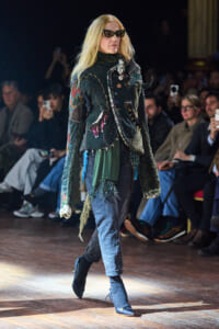 Model walking a fashion show in a patchwork, fringed jacket, jeans, blue heels, and sunglasses on the catwalk.