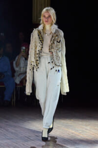 Model walking a fashion runway in a pale, embellished jacket layered over a cream turtleneck and light gray sweatpants, accessorized with a beaded necklace and a crown-like headpiece.