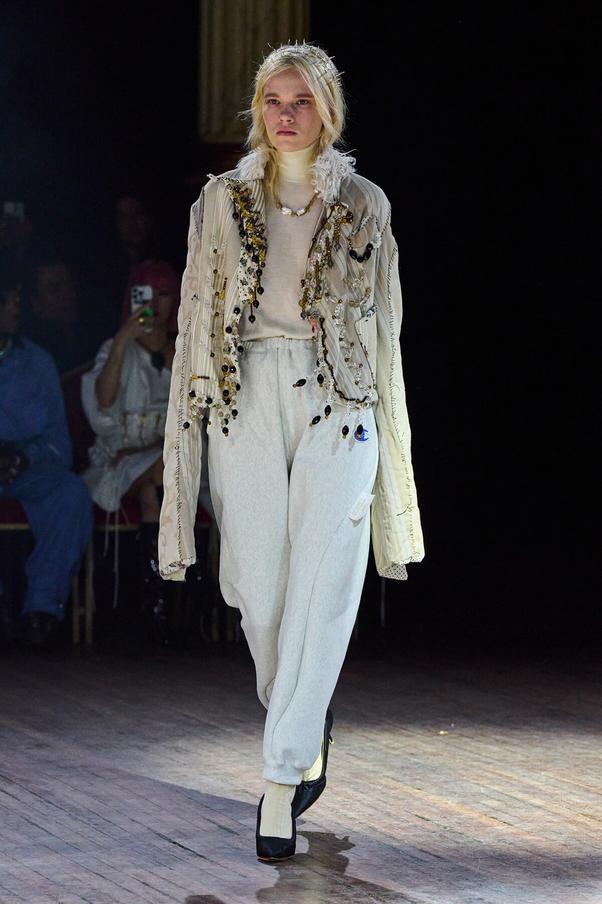 Model walking a fashion runway in a pale, embellished jacket layered over a cream turtleneck and light gray sweatpants, accessorized with a beaded necklace and a crown-like headpiece.