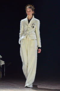 Model on runway wearing a white textured suit with a double-breasted blazer and wide-leg pants, gold spike headpiece