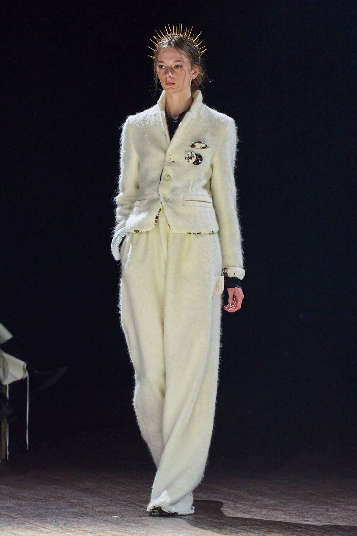 Model on runway wearing a white textured suit with a double-breasted blazer and wide-leg pants, gold spike headpiece
