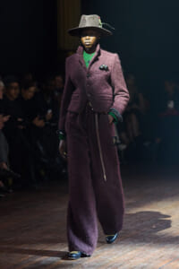 Model walking a fashion runway in a deep plum textured suit with wide legs, green top, and brown hat with a decorative pin set.