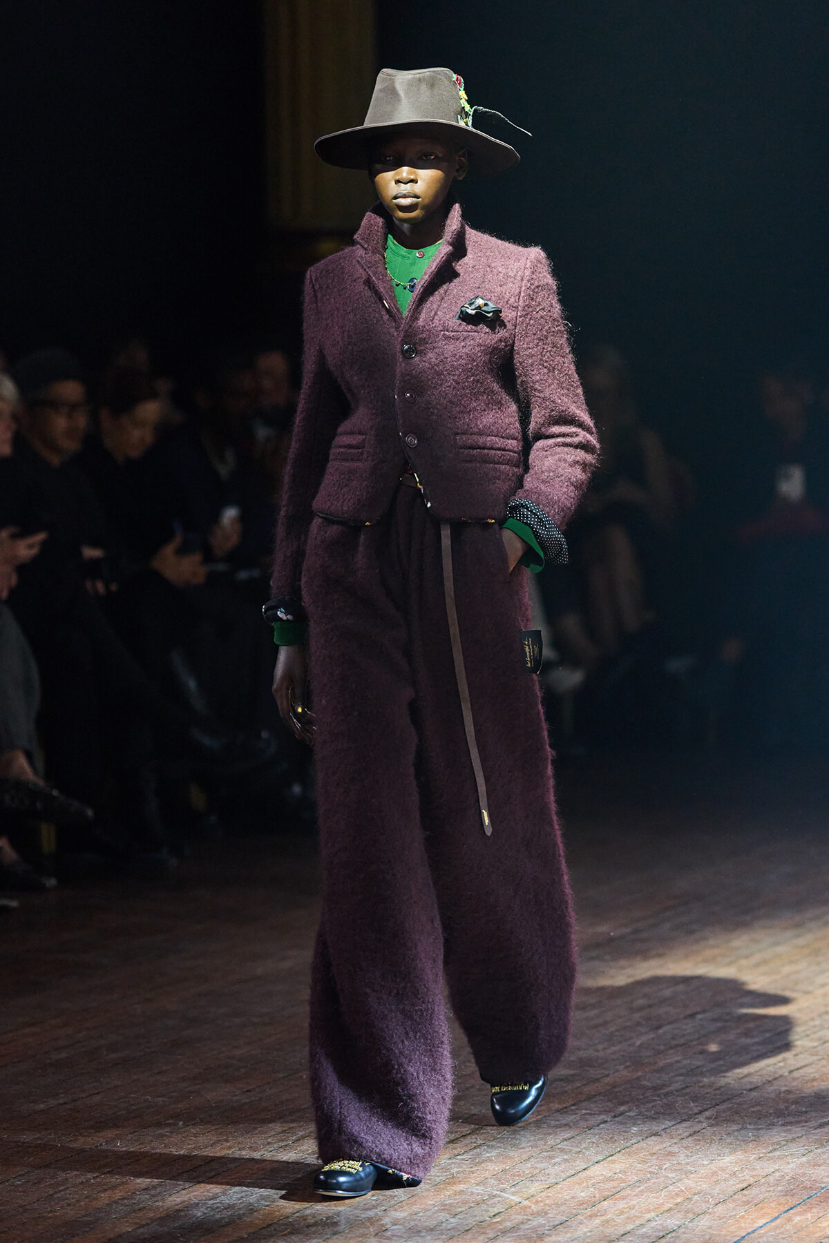 Model walking a fashion runway in a deep plum textured suit with wide legs, green top, and brown hat with a decorative pin set.