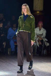 Model on a runway wearing an olive-green cardigan over a navy shirt with brown wide-leg pants and black loafers with gold embroidery.