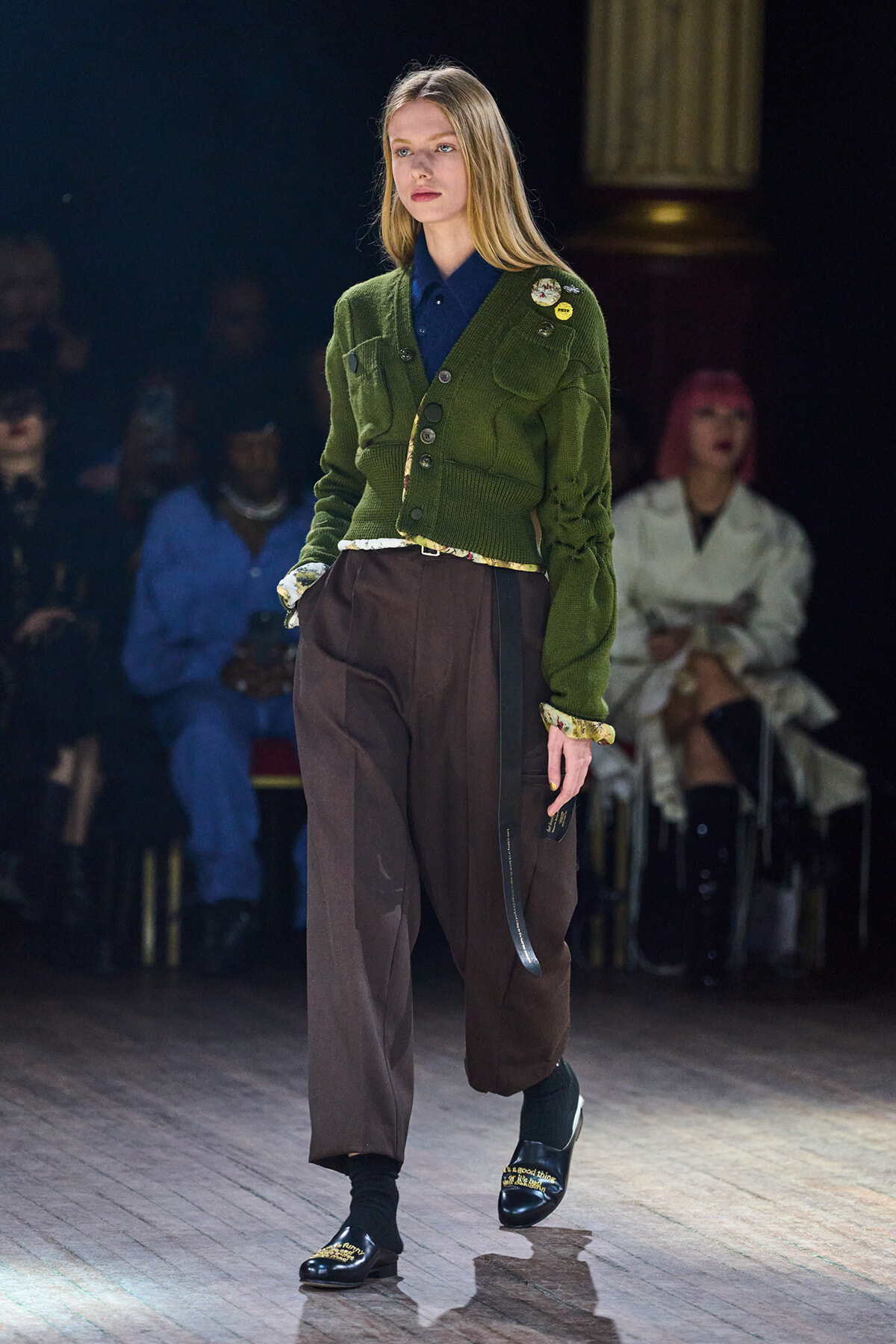 Model on a runway wearing an olive-green cardigan over a navy shirt with brown wide-leg pants and black loafers with gold embroidery.