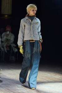Model walking a runway in a light denim jacket, blue wide-leg jeans, a fringe scarf, and a gold glove on one hand.