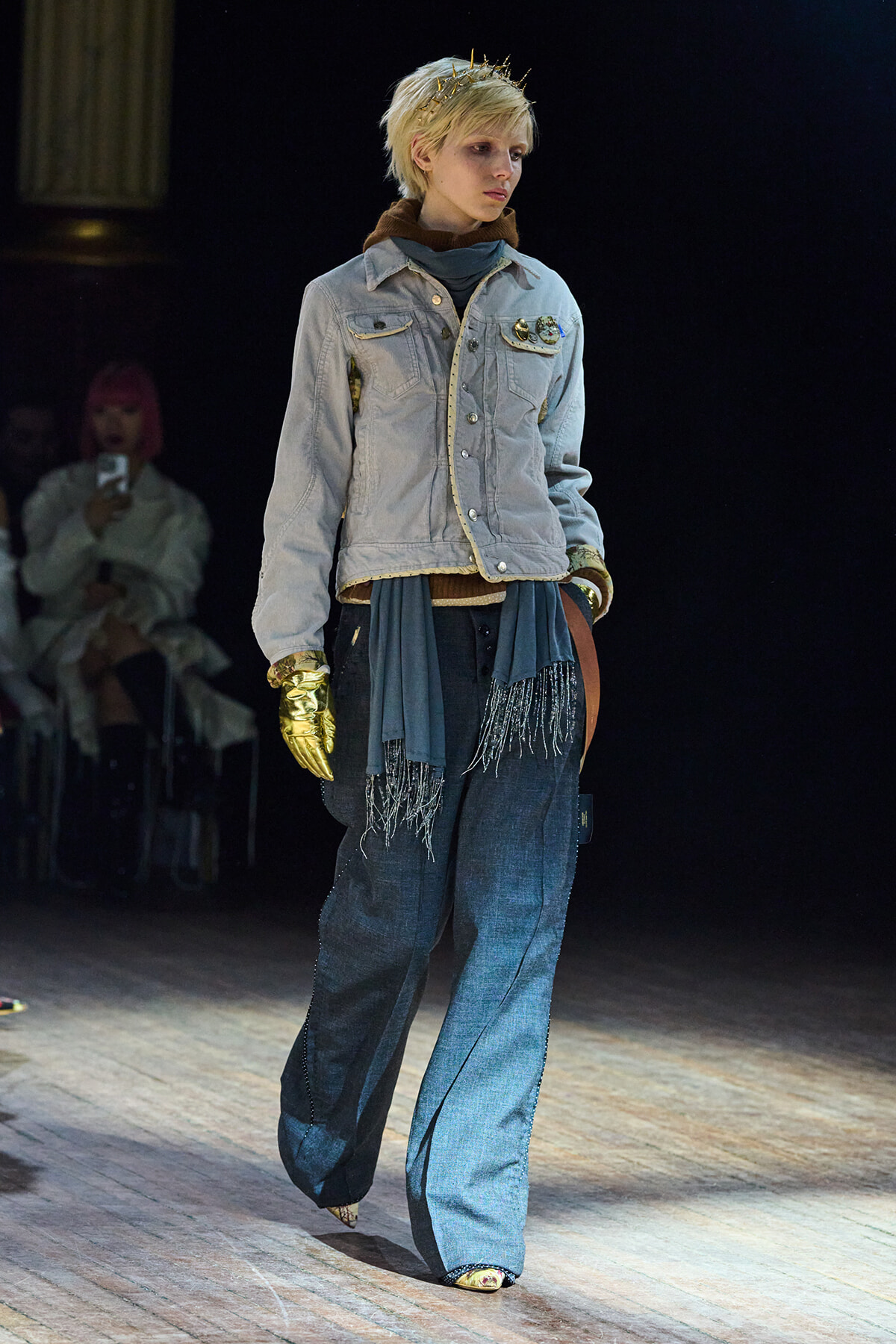 Model walking a runway in a light denim jacket, blue wide-leg jeans, a fringe scarf, and a gold glove on one hand.