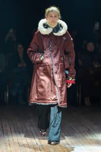 Model walking a fashion show in a burgundy knee-length coat with a large white fluffy scarf and patterned shoes on the catwalk.