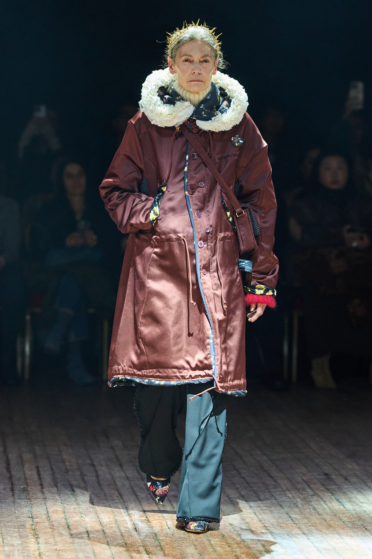 Model walking a fashion show in a burgundy knee-length coat with a large white fluffy scarf and patterned shoes on the catwalk.