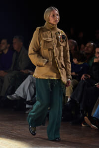 Runway model wearing a tan military-style jacket with medals and teal wide-leg pants on a wooden catwalk, audience in the background.