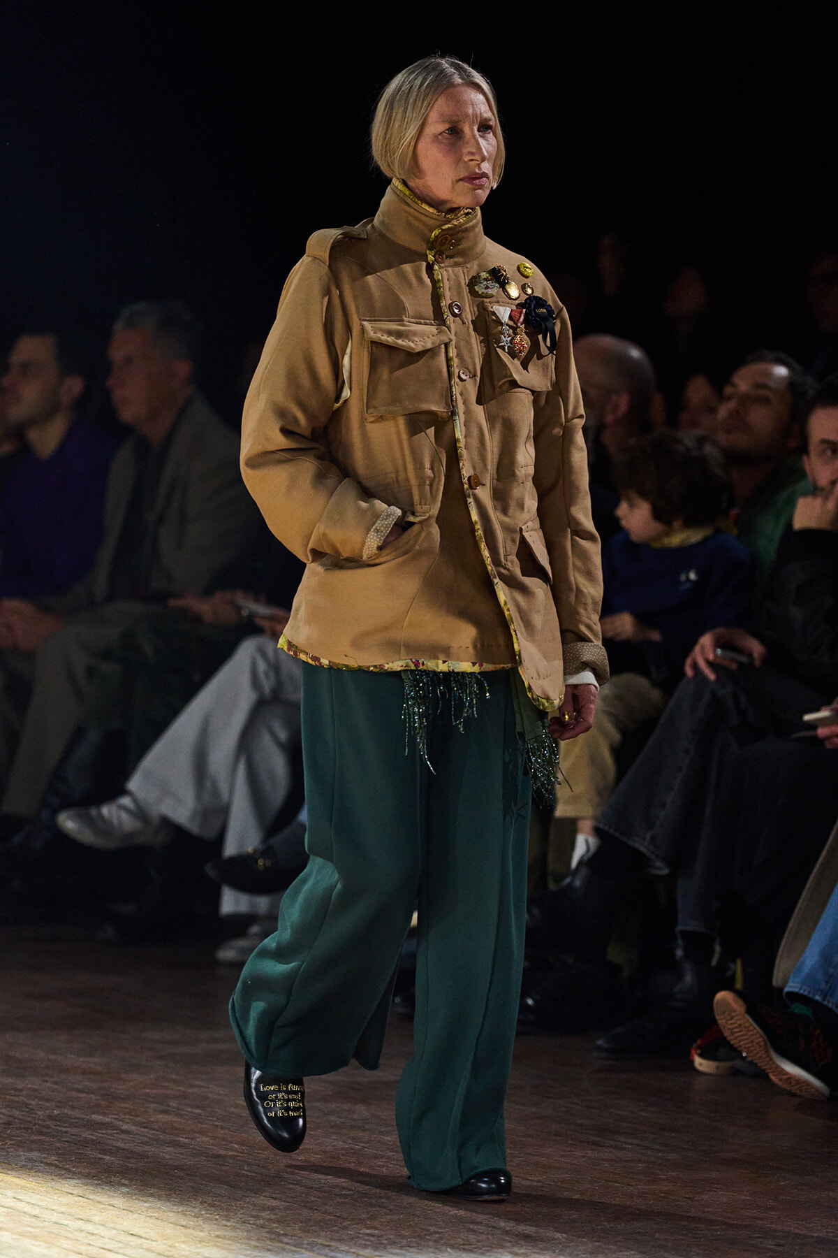 Runway model wearing a tan military-style jacket with medals and teal wide-leg pants on a wooden catwalk, audience in the background.