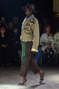 Model walks a runway in a beige pin-covered jacket, green scarf, brown cropped trousers, and floral heels with a wide-brimmed hat.