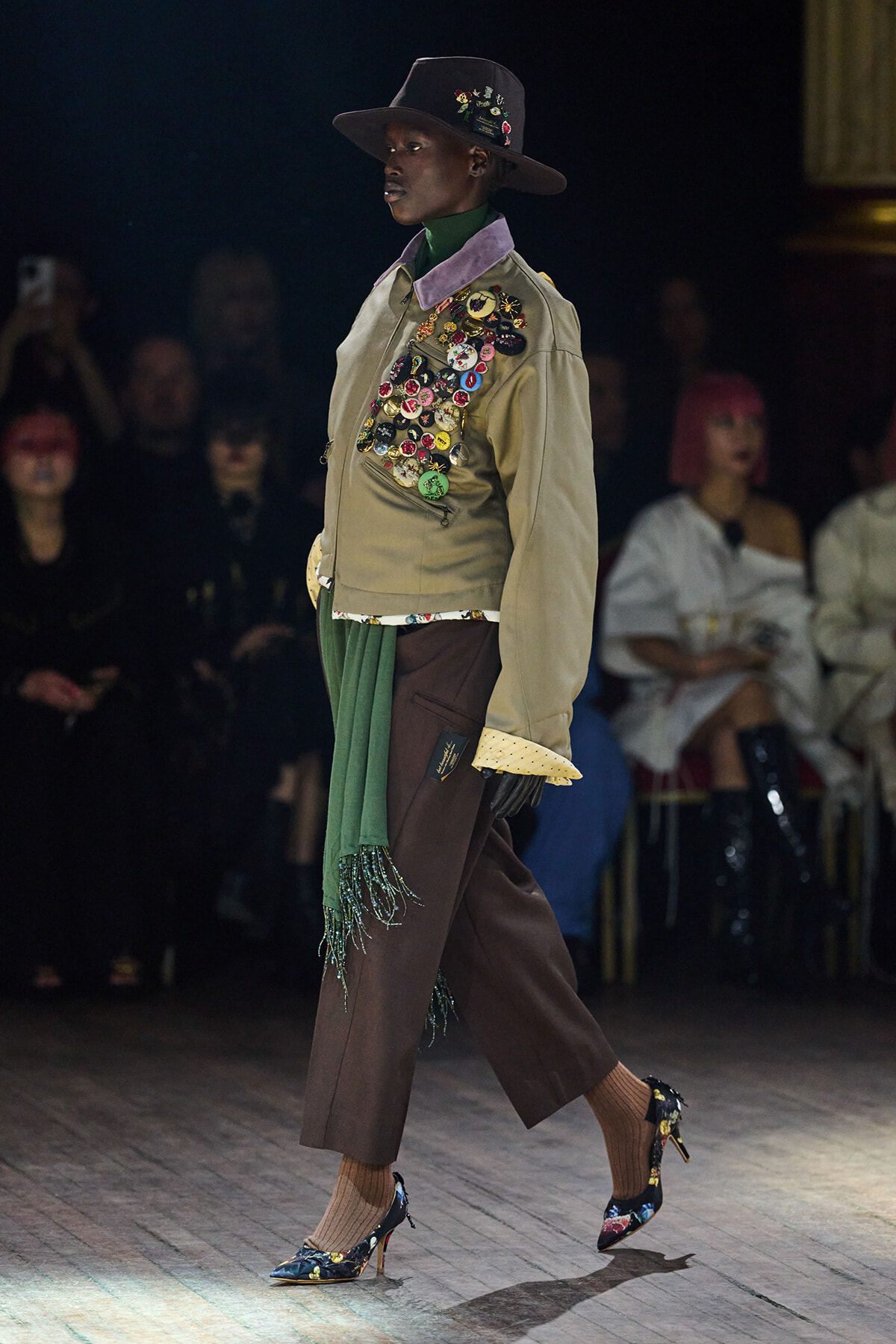 Model walks a runway in a beige pin-covered jacket, green scarf, brown cropped trousers, and floral heels with a wide-brimmed hat.