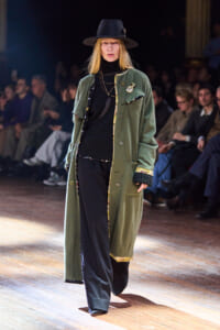 Model walking runway in an oversized olive-green coat over a black turtleneck, layered necklaces, and a wide-brimmed hat.