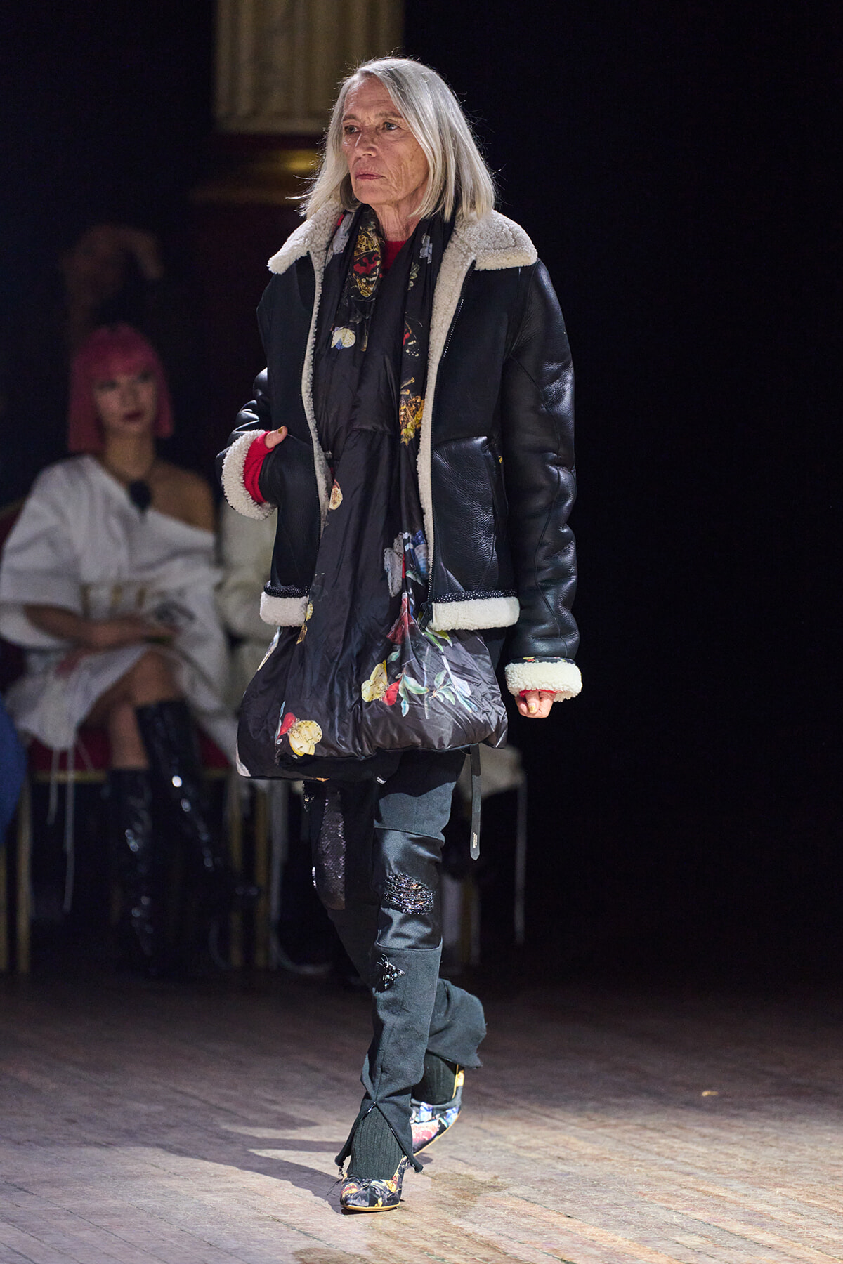 Older woman walking a fashion runway in a black shearling-lined jacket, scarf, and floral tote; focused expression.