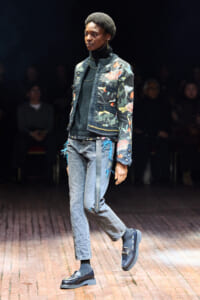 Model walking a fashion runway in a floral-embroidered denim jacket, black turtleneck, gray jeans, and blue loafers.